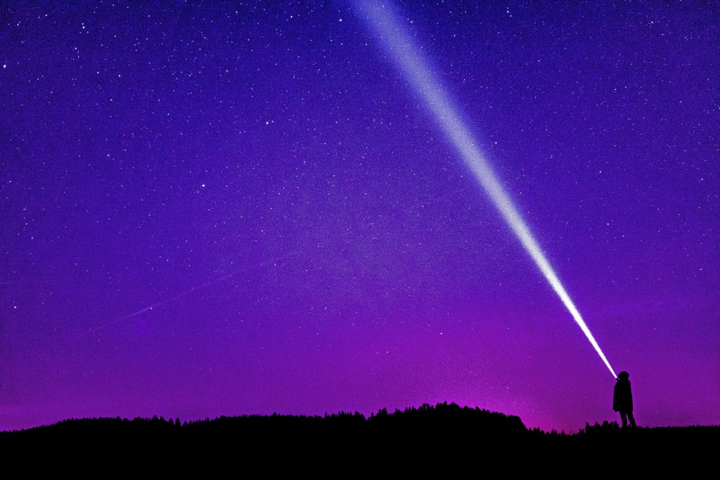 A flat black landscape against a bright pink-to-purple sunset background. Someone in the foreground looks up into space as a silhouette, a bright white beam of light stretching into the stars from their eyes.