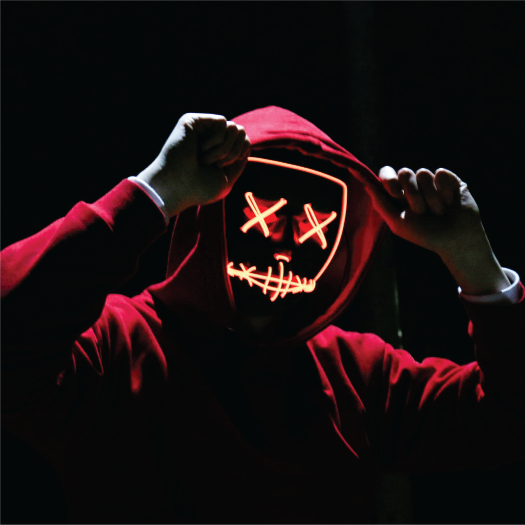 Human in a red hoodie against a black, shadowed background holds their fists up by their temples, their face obscured by a glowing orange LED mask with Xs for eyes and a jagged line for the mouth.