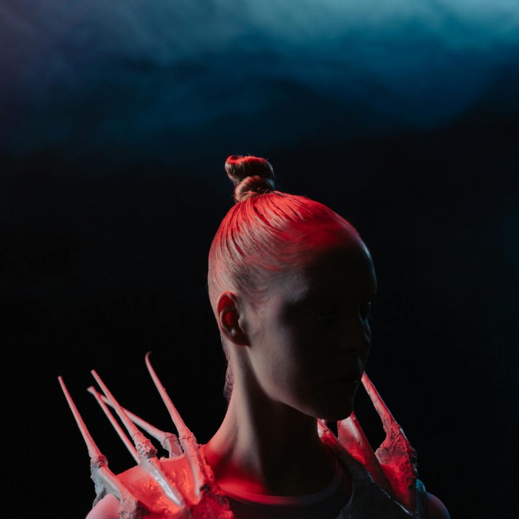 A human stands in shadow, their face almost complete obscured against the dark backdrop, with bright pink hair pulled into a tight, thin bun and spikes protruding from their shoulders.