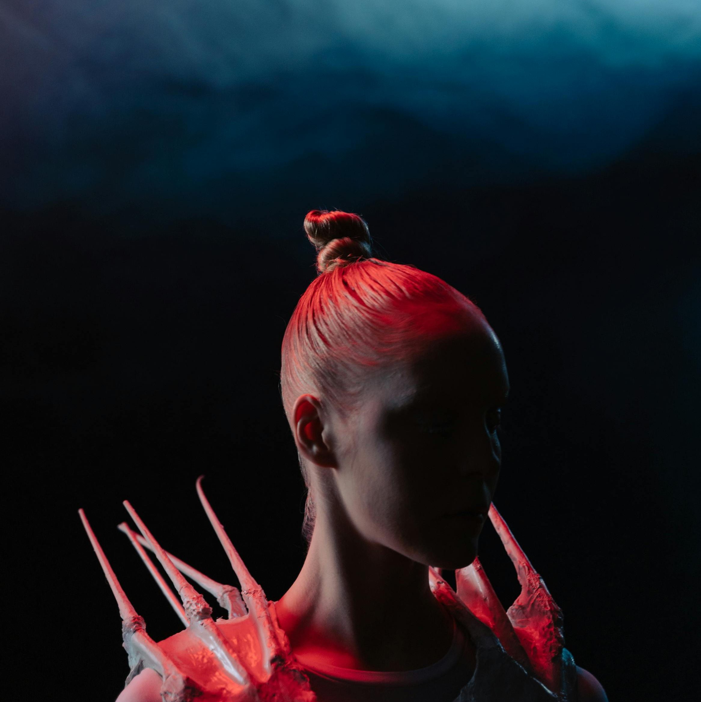 A human stands in shadow, their face almost complete obscured against the dark backdrop, with bright pink hair pulled into a tight, thin bun and spikes protruding from their shoulders. The image is meant to depict a Human spacer preparing their Rapadruk costume for the Starmourn sector. 