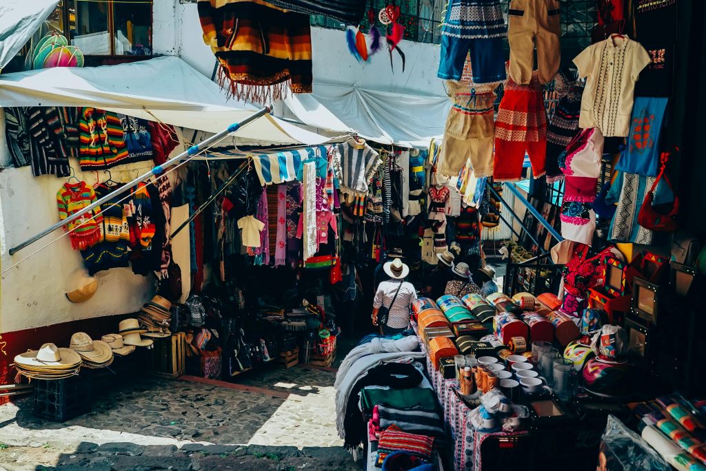 Colorful clothing lines the walls of a packed vendor stall, shirts, blankets, hats and more displayed across a collection of tables and hanging from the rafters as a customer stands in the center, looking tiny amidst all the offerings. The image is meant to capture an example of a cluttered stall in the Starmourn Floating Market. 