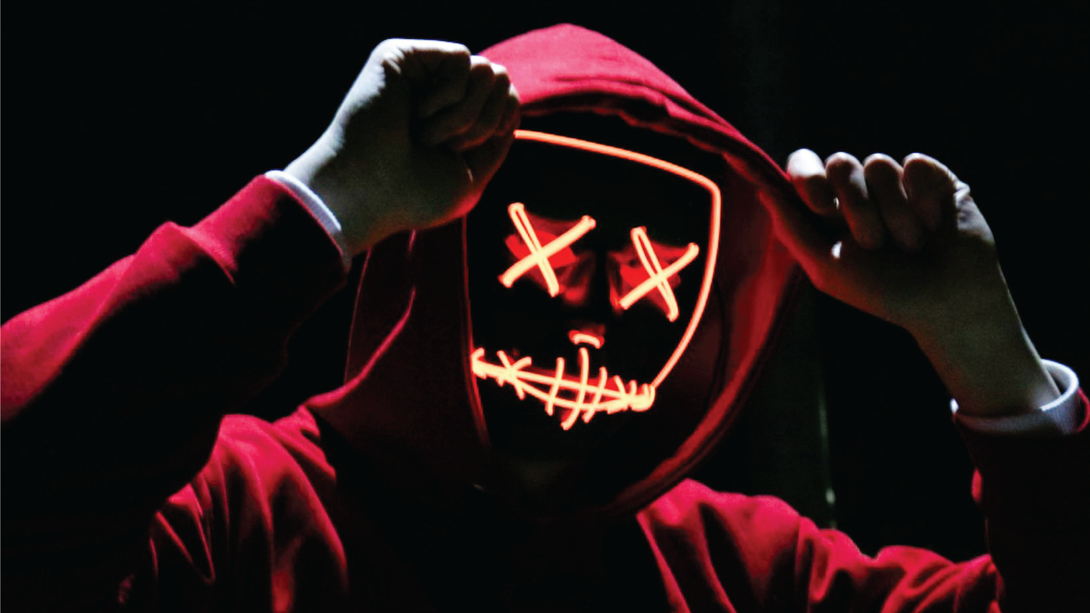 Human in a red hoodie against a black, shadowed background holds their fists up by their temples, their face obscured by a glowing orange LED mask with Xs for eyes and a jagged line for the mouth.